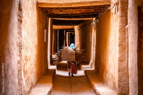 Woman carrying a load of dry palm leaves through the narrow alleyway in El Khorbat, Morocco