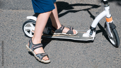 Child's feet in sandals standing on the white scooter in motion. black scooter wheels. 