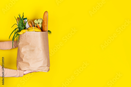 Girl or woman holds a paper bag filled with groceries such as fruits, vegetables, milk, yogurt, eggs isolated on yellow.