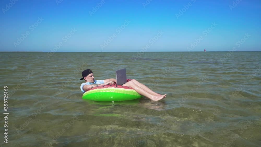 A young business person is enthusiastically working behind a laptop swaying on the waves of sea sitting on a inflatable circle. 4K A young business person is enthusiastically working behind a laptop swaying on the waves of sea sitting on a inflatable circle. 4K