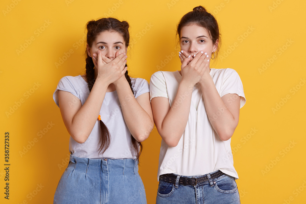 Attractive young females in white t shirts and jeans, posing against yellow wall and covering mouths with palms, looking directly at camera with big eyes, see something shocking. Attractive young females in white t shirts and jeans, posing against yellow wall and covering mouths with palms, looking directly at camera with big eyes, see something shocking.
