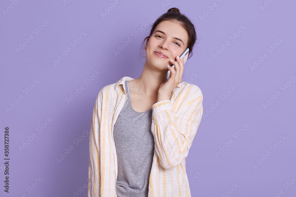 Satisfied woman talking via modern smart phone and looking directly at camera, looks happy, being in good mood, stands against lilac background, girl with pleasant appearance. Satisfied woman talking via modern smart phone and looking directly at camera, looks happy, being in good mood, stands against lilac background, girl with pleasant appearance.