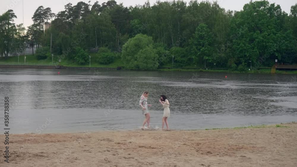 Two young caucasian girls in summer dresses play and dance in rain in pond of city park. Girls are free froom studying and work. Two young caucasian girls in summer dresses play and dance in rain in pond of city park. Girls are free froom studying and work.