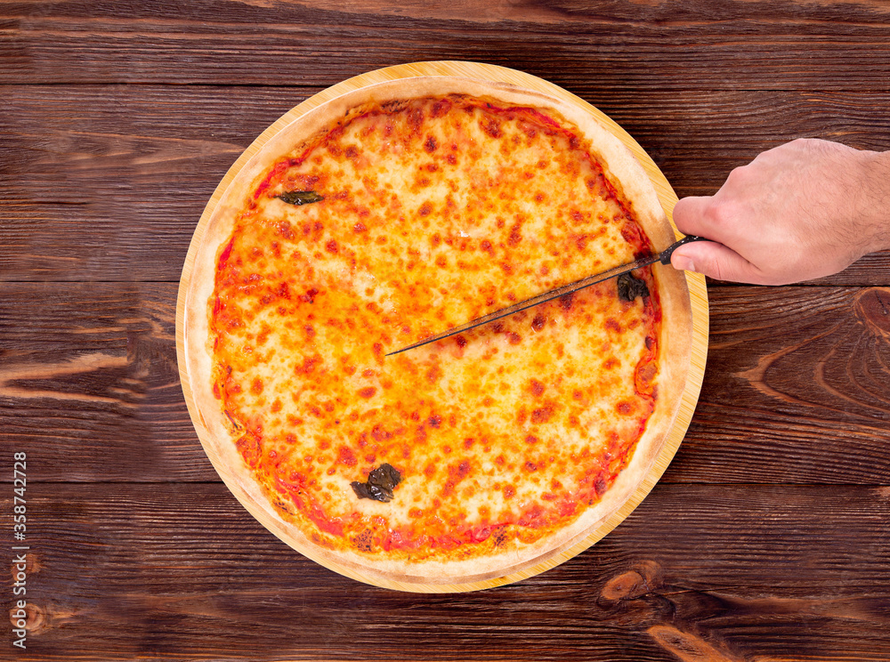 Top view of male hand which cutting the pizza with a kitchen knife. Delicious pizza with mozzarella, bocconcini and basil leaves or pizza margherita on a wood platter which is on wooden rustic table Top view of male hand which cutting the pizza with a kitchen knife. Delicious pizza with mozzarella, bocconcini and basil leaves or pizza margherita on a wood platter which is on wooden rustic table
