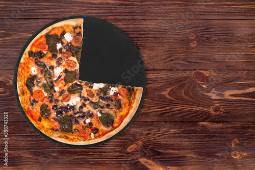 Delicious pizza with cherry tomatoes, spinach, mozzarella, feta, kalamata olive and mushrooms, without a quarter on round slate platter which is on wooden table, top view and space for text