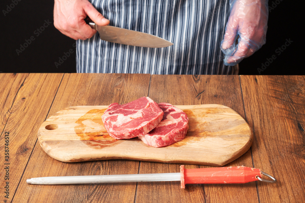 Two fresh raw rib eye steaks on a wooden cutting board, Premium range of beef. Butcher in black and white apron holding knife. Black background. Steel with red handle in foreground.
