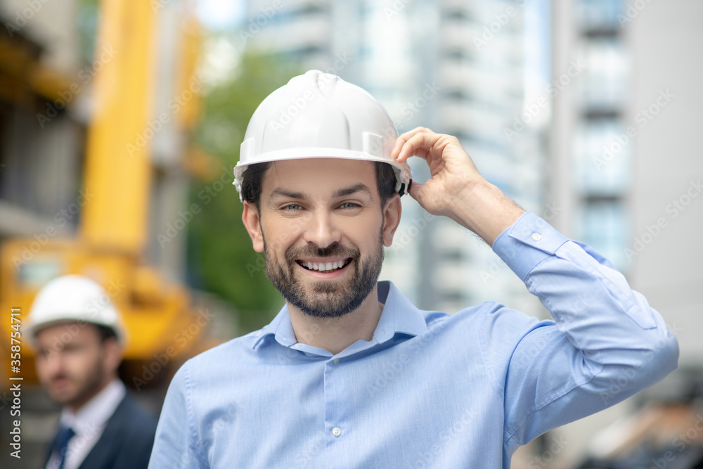 Foreman holding his helmet with one hand, smiling, building supervisor standing behind him Foreman holding his helmet with one hand, smiling, building supervisor standing behind him