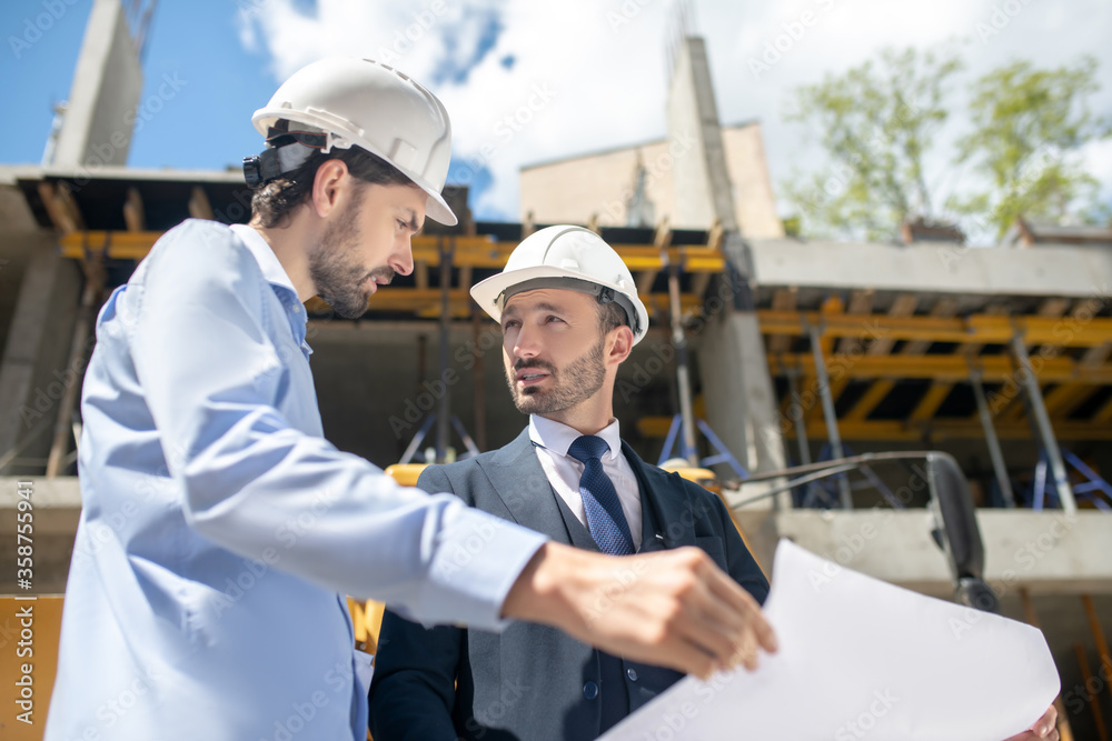 Building supervisor and foreman standing in front of building, discussing and checking blueprint Building supervisor and foreman standing in front of building, discussing and checking blueprint
