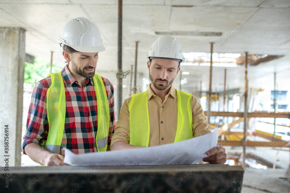 Building workers brainstorming over blueprint, discussing it Building workers brainstorming over blueprint, discussing it