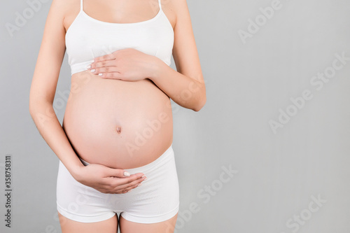Cropped image of pregnant woman in white underwear holding her belly at gray background. Future mother is waiting her baby. Copy space