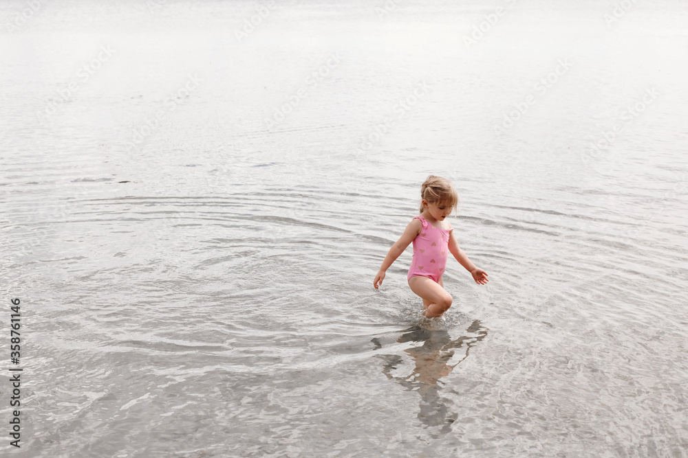 A three-year-old girl of European appearance plays in the water on the beach. A child walks ankle-deep in the lake. Holidays in nature. Summer photo of a baby in a swimsuit. A three-year-old girl of European appearance plays in the water on the beach. A child walks ankle-deep in the lake. Holidays in nature. Summer photo of a baby in a swimsuit.