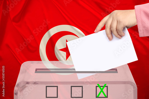female voter lowers the ballot in a transparent ballot box against the background of the national flag of Tunisia, concept of state elections, referendum photo