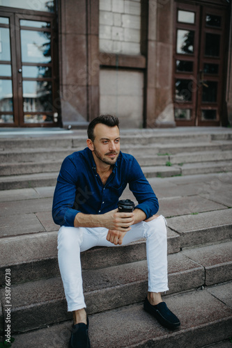 Young man sits and dreams on stairs with cup of coffee near building.