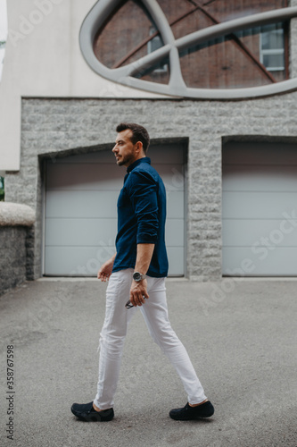 Young man goes at street with sunglasses in his hands near building.