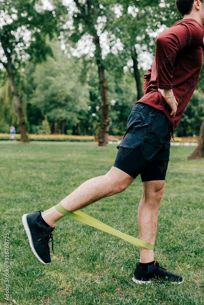 Fototapeta premium Cropped view of sportsman using elastics band while training on lawn in park