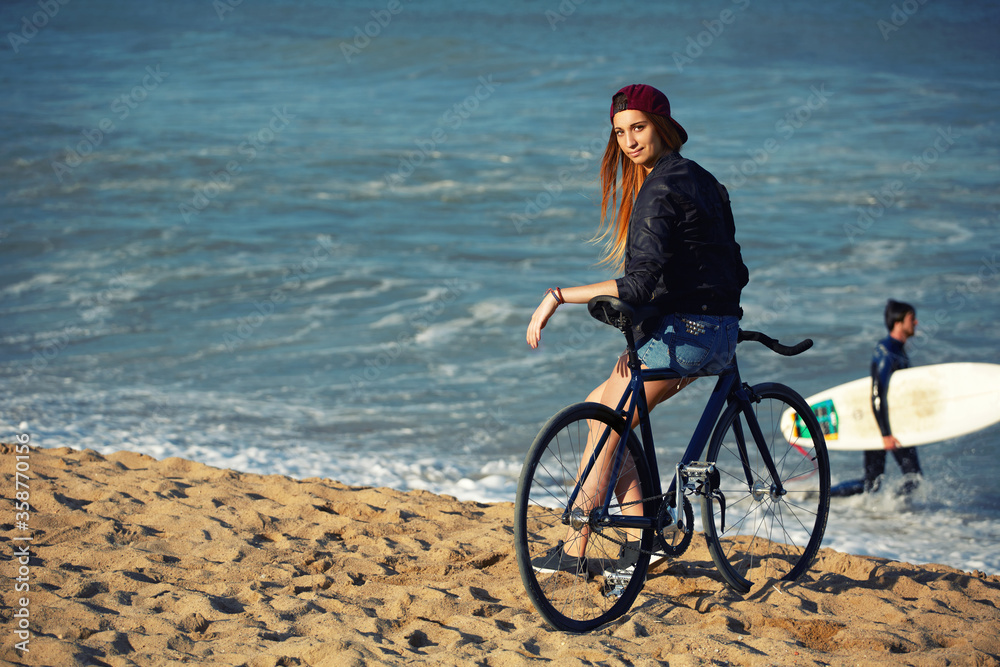 Attractive hipster girl relaxing on the beach after ride on her sport fixed gear bicycle, young woman standing on seashore enjoying sunny afternoon, charming female with rental bike outdoors, filter