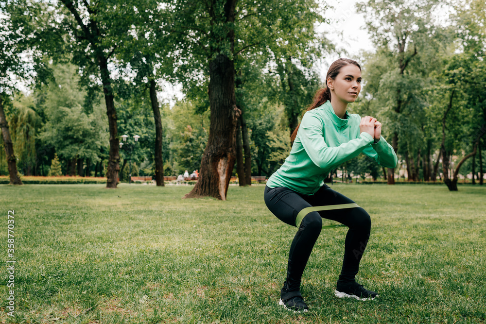 Attractive sportswoman doing squat with resistance band in park Attractive sportswoman doing squat with resistance band in park