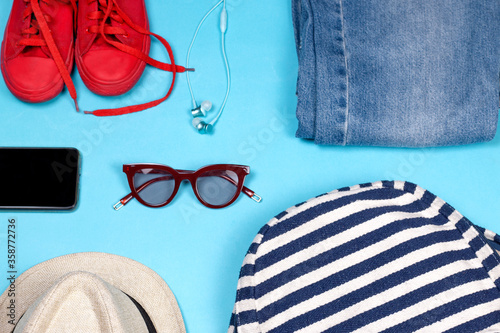 Journey and holiday vacation image with travel bag, Jeanes, red sneakers, sunglass, Headphones, phone and summes hat on blue background.