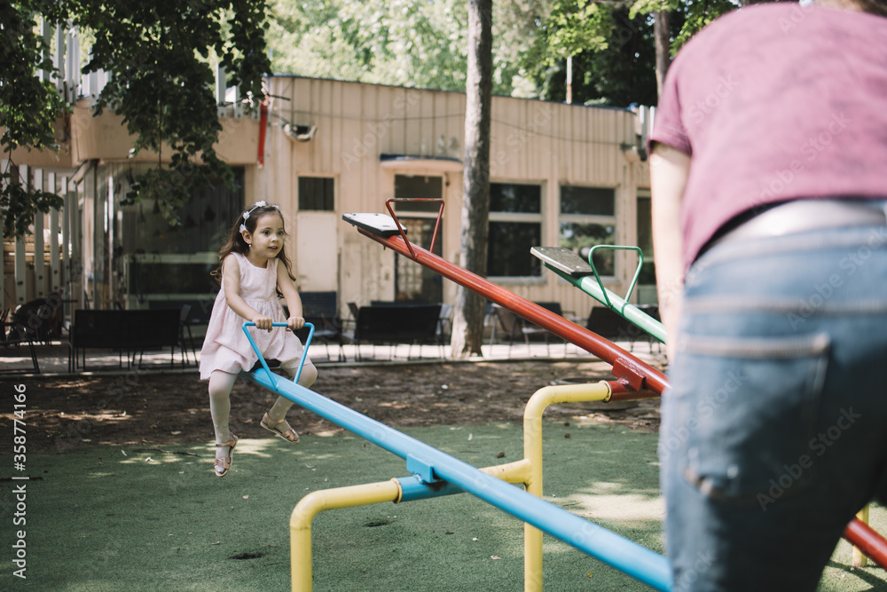Fototapeta premium Cute little girl with headband sitting on seesaw. Back of man playing with little girl on seesaw in park.