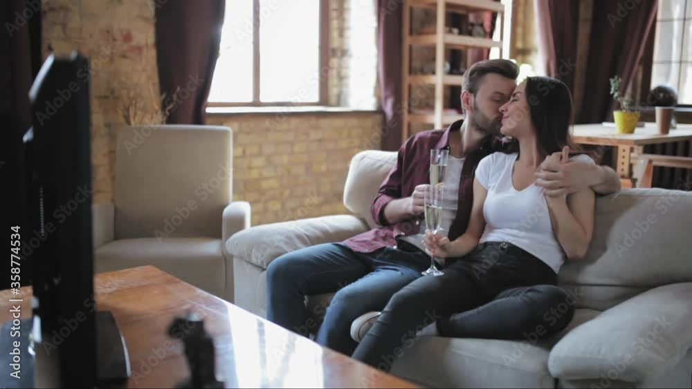 Young people celebrates anniversary having a glass of champagne while sitting on a couch in a living room in front of tv set watching their wedding day. Family concept. Prores 422.