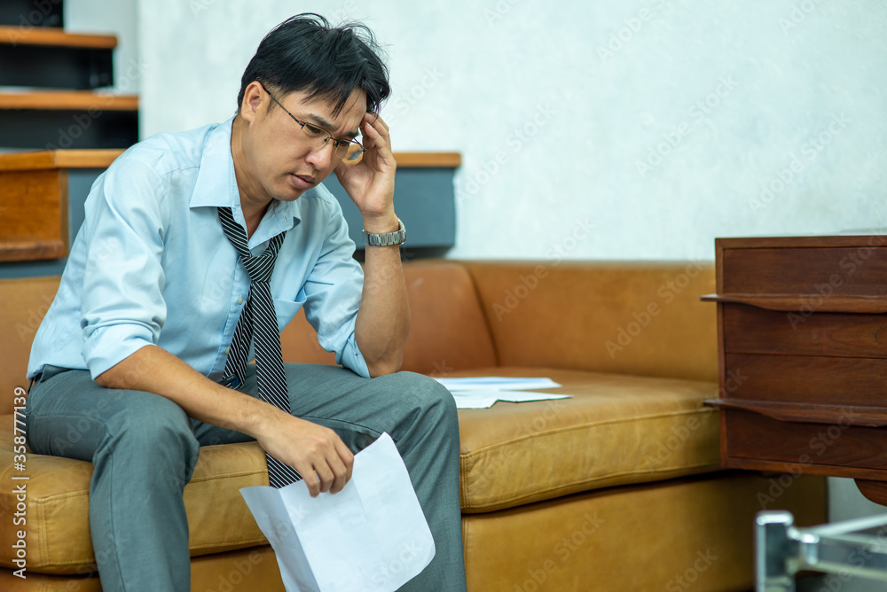 Stressed Asian businessman sitting on sofa with holding financial bill in hand and think of debt problem. Entrepreneur man worry about money management. Mental health and economic problem concept