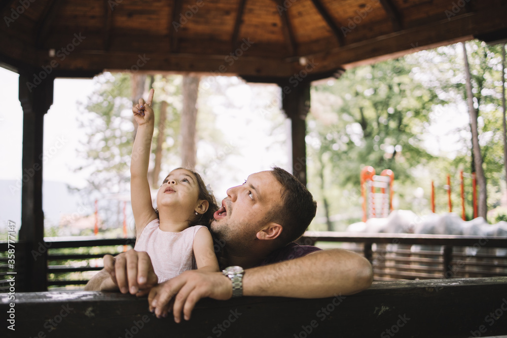 Daughter and father having fun in wooden gazebo. Little girl hand pointing while standing with her dad in wooden pavilion in nature.