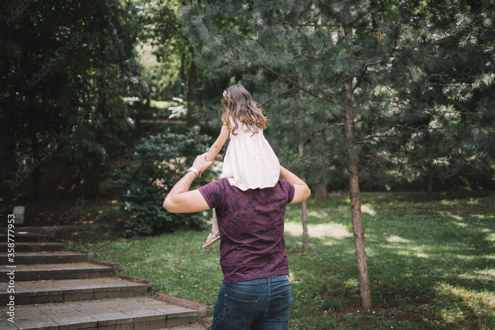 Naklejka premium Back view of father having daughter on his shoulders. Dad carrying his little girl on shoulders while walking in forest.