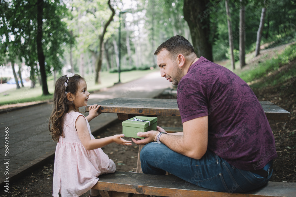 Dad and little girl sitting on wooden bench with present. Side view of daughter and father holding present while sitting on bench in park. Dad and little girl sitting on wooden bench with present. Side view of daughter and father holding present while sitting on bench in park.