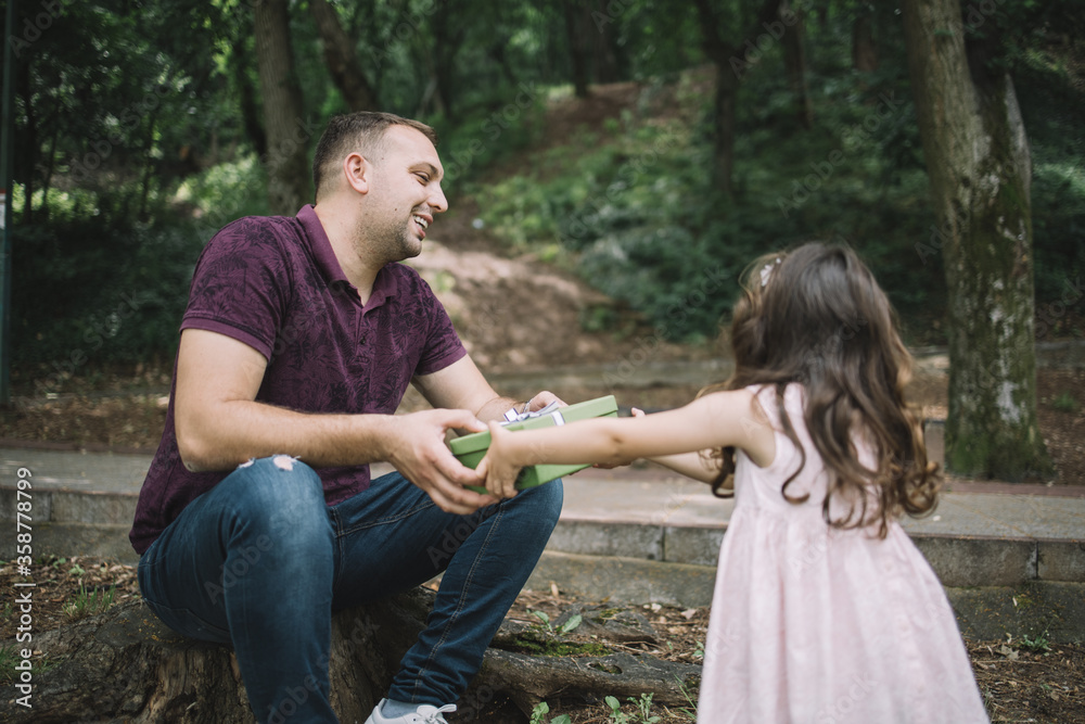 Fototapeta premium Dad and small girl holding present in park. Father sitting on stump in park giving present to his little daughter.