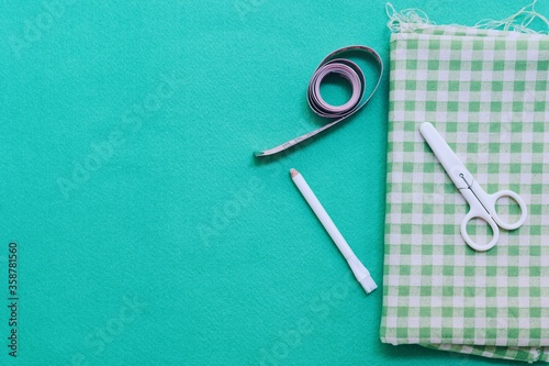 Close-up view of folded Gingham fabric with accessories like scissor  tape measure and fabric marker on solid color felt background. Top view  copy space ideas.