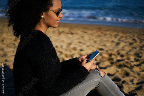 Attractive afro american female recreating sitting on beach admire sea view using smartphone to chatting with followers in social network updating profile and sharing multimedia files from trip