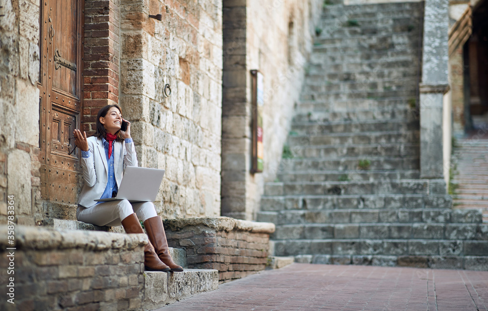 young female talking on her cell phone with laptop, sitting outdoor on stairways in italy