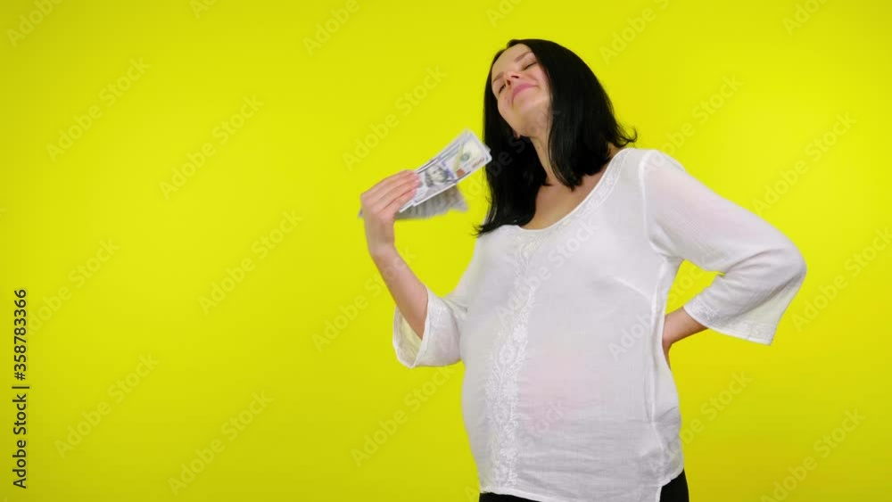 Rich young pregnant woman with black hair in a white blouse waves money in front of face on a yellow background with copy space. Place for text or product. 4K slow motion footage Rich young pregnant woman with black hair in a white blouse waves money in front of face on a yellow background with copy space. Place for text or product. 4K slow motion footage