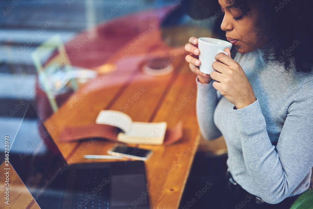 Cropped image of attractive female with curly hair enjoying tasty beverage during work break in coffee shop.Pretty hipster girl spending leisure time with aroma drink sitting at table with gadget. Cropped image of attractive female with curly hair enjoying tasty beverage during work break in coffee shop.Pretty hipster girl spending leisure time with aroma drink sitting at table with gadget.