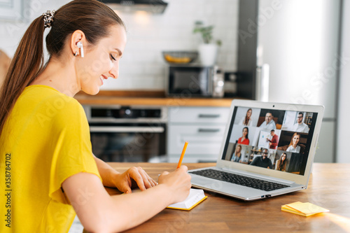 Side view of young woman watching on laptop display with a group of multiracial people on it and writing notes. Video call, online conference, webinar photo