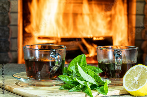 Tea with lemon and mint in glass cups in front of burning fireplace in a country house.