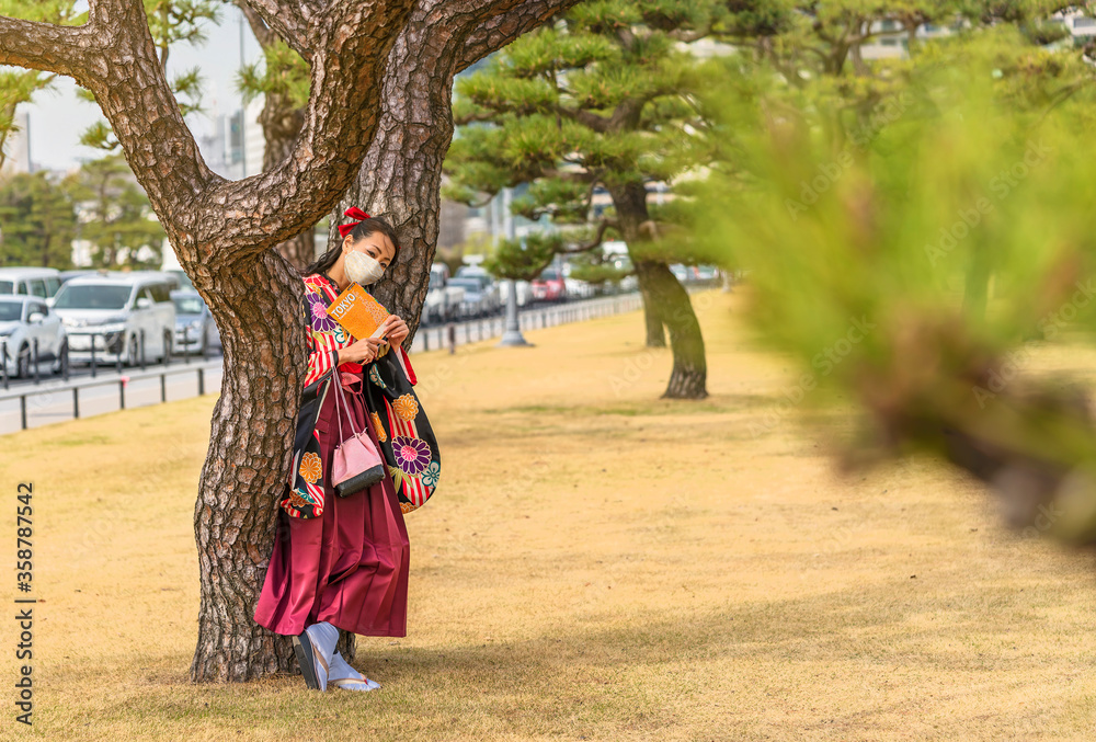 Japanese woman dressed in a hakama kimono and wearing a facial mask reading a Tokyo travel guide book leaned against the trunk of pine tree in the Kokyo Gaien National Garden of Tokyo Imperial Palace Japanese woman dressed in a hakama kimono and wearing a facial mask reading a Tokyo travel guide book leaned against the trunk of pine tree in the Kokyo Gaien National Garden of Tokyo Imperial Palace