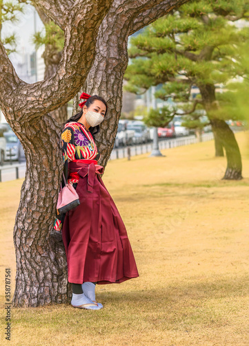 Japanese woman dressed in a hakama kimono and wearing a facial mask leaned against the trunk of a pine tree in the Tokyo Imperial Palace.