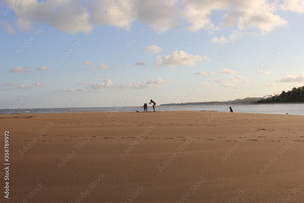 Guarajuba beach, Bahia, Brazil. Wide and open beach with beige sand at sunset. In the middle of the beach a man and two children. In the background, ocean water and vegetation.