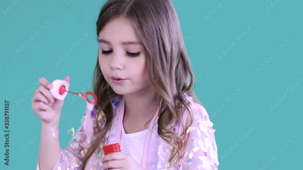 An attractive little girl blows some soap bubbles at the camera at studio on blue background. Concept of childhood and joy. An attractive little girl blows some soap bubbles at the camera at studio on blue background. Concept of childhood and joy.