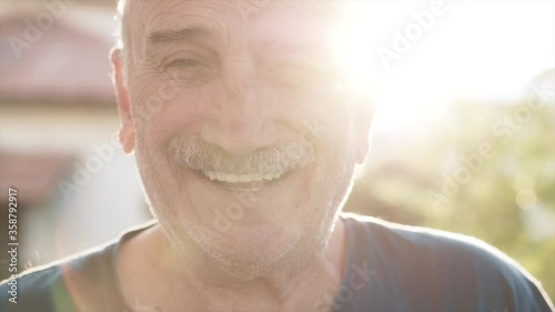 Sympathetic old senior man's face portrait. Happy retired elderly guy is smiling to camera in afternoon time, with sun flare