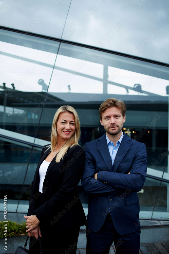 Two business colleagues standing against office building with brightly smile, experienced businessman standing with his female colleague smiling and looking to you, teamwork concept Two business colleagues standing against office building with brightly smile, experienced businessman standing with his female colleague smiling and looking to you, teamwork concept