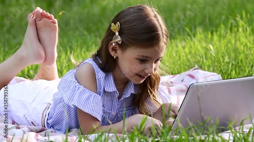 Caucasian pretty child with long hair looking at the laptop, lying on the blanket, texting email and watching at the camera. Concept of lifestyle and childhood.