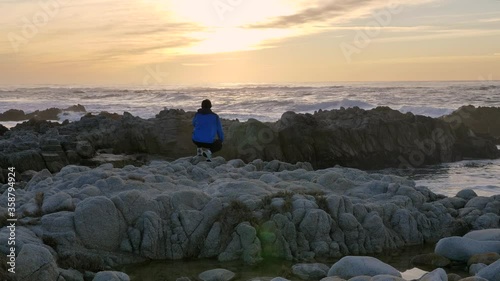 Slow motion cinematic of man sitting in blue jacket and walking at rocky coast at sunset california vibes pacific ocean