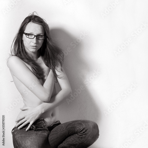 Sexy young woman wearing only jeans and glasses, she is hiding her breasts wirh her arms, monochrome photo