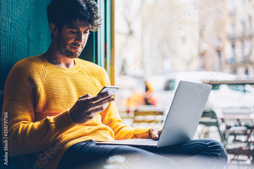 Young man reading sms message from friend on smartphone sitting with laptop device in stylish coworking space.Skilled student installing new application on mobile phone via free internet connection