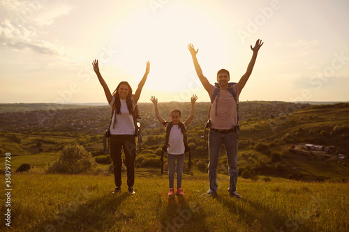 Young parents and their kid with backpacks raising hands on mountain top at sunset. Happy family on summer camping trip