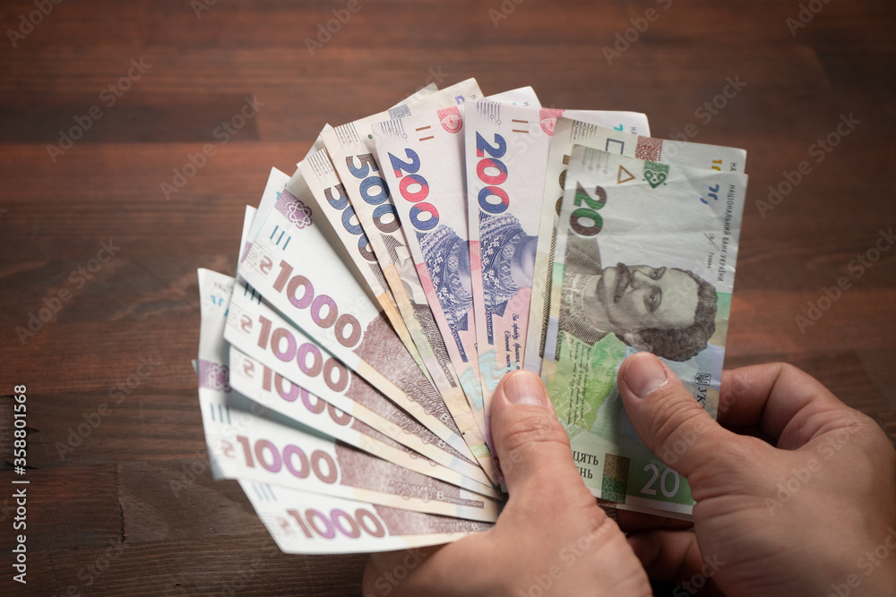 Male hands holding banknote fan. Caucasian man has pack of new Ukrainian hryvnias against of wooden table background. Close up shot. Cash money concept.