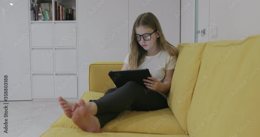 Barefoot girl in glasses sitting on sofa and watching video on tablet while resting at home during quarantine