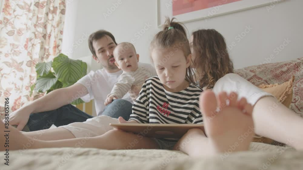 A Young Family, Father, Mother and Two Baby Daughters, a Preschooler and an Infant, are Having Fun and Use Tablet Computer, Sitting on the Bed. Cozy White Bedroom Interior. A Young Family, Father, Mother and Two Baby Daughters, a Preschooler and an Infant, are Having Fun and Use Tablet Computer, Sitting on the Bed. Cozy White Bedroom Interior.